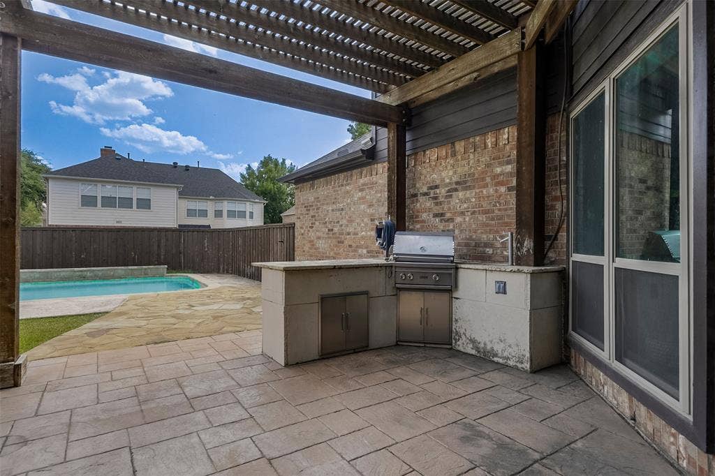1200 Lakeway Drive, Plano, TX 75075 Expansive patio with built-in outdoor kitchen featuring stainless steel appliances and stone countertop, complemented by a pergola overhead and inground swimming pool visible beyond