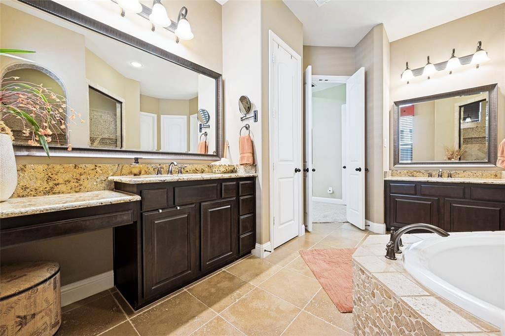 This luxurious primary bathroom features dual vanities with granite countertops, generous cabinetry, large framed mirrors, a linen closet, and a built-in soaking tub.