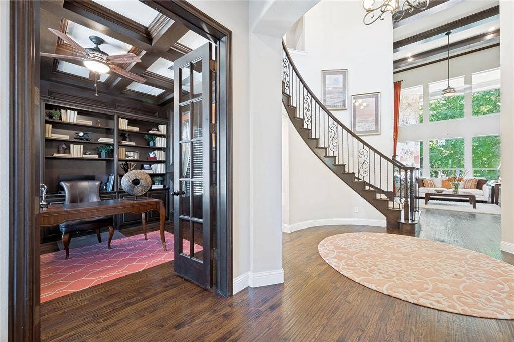 The residence features a stunning entryway distinguished by a grand staircase and elegant hardwood flooring. The adjacent office is appointed with French doors, a coffered ceiling, and custom built-in shelving.