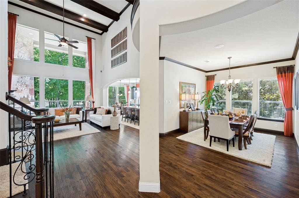 Upon entering the home, you are welcomed into a two-story living area featuring expansive windows, hardwood flooring, wood-beamed ceilings, and an elegant decorative staircase.