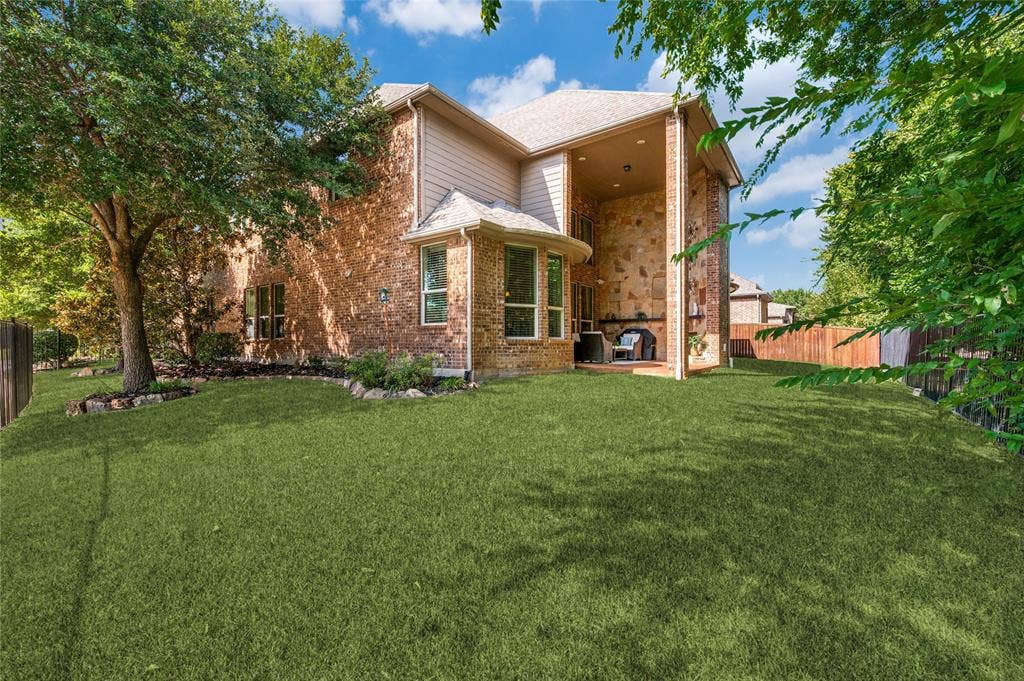 This lush backyard features a well-manicured lawn, mature trees, and professional stone-edge landscaping. The space includes a covered patio equipped with a stone fireplace, perfect for outdoor entertaining.