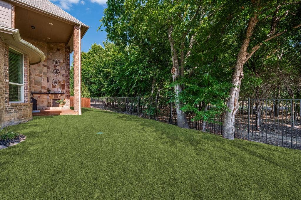 Two-story stone-walled covered patio with fireplace and rear yard featuring a black wrought iron fence, landscaping and mature trees--perfect for entertaining or relaxing.