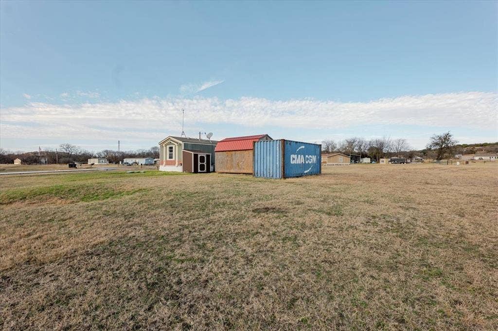 126 Starry Sky Court, Springtown, TX 76082 Open field with a small structure featuring light-colored siding and a dark roof, accompanied by a blue shipping container and a red-roofed shed