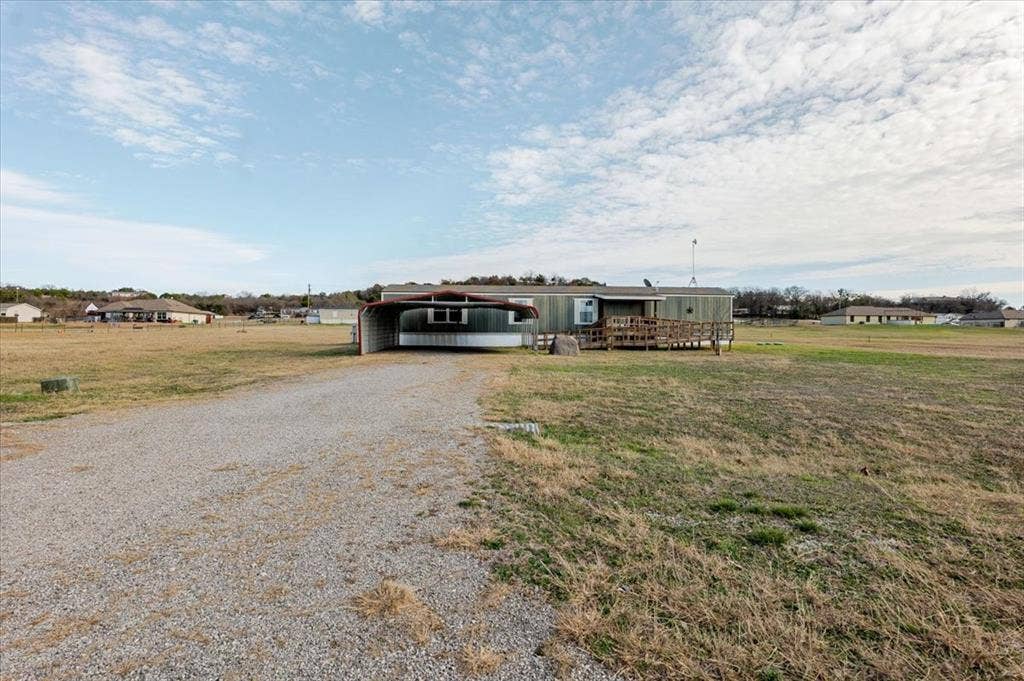 126 Starry Sky Court, Springtown, TX 76082 Expansive gravel driveway leading to a residence featuring a covered carport and an exterior deck with a ramp