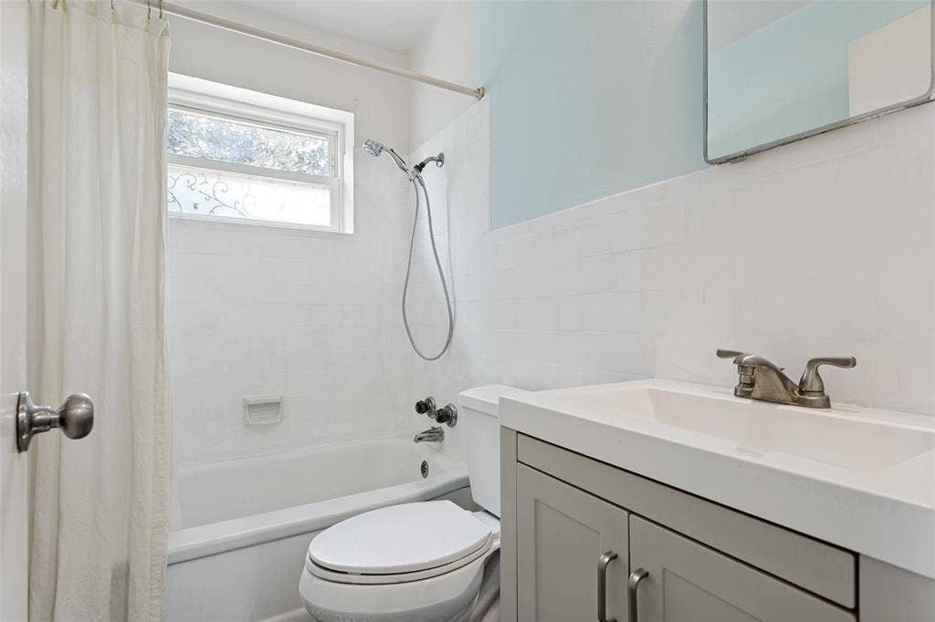 2312 Brooks Street, Garland, TX 75041 Bathroom featuring a white bathtub, tiled shower surround, window with privacy glass, single-basin vanity with gray cabinetry, and a toilet