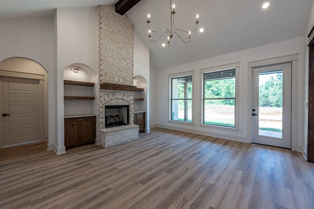 17049 Island Lake Drive, Tyler, TX 75708 Bright open-concept living area featuring wood-finish flooring throughout, a striking stone fireplace with built-in shelving and lower cabinetry, arched architectural details, and abundant natural light from triple glass doors and multiple windows
