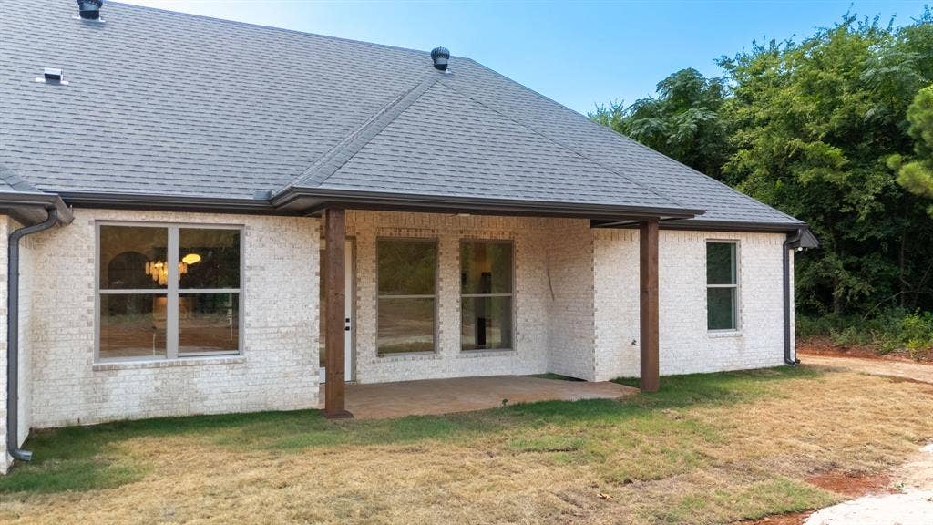 17049 Island Lake Drive, Tyler, TX 75708 Rear exterior featuring a covered concrete patio supported by wood-finish pillars, white brick façade, and a dark shingle roof