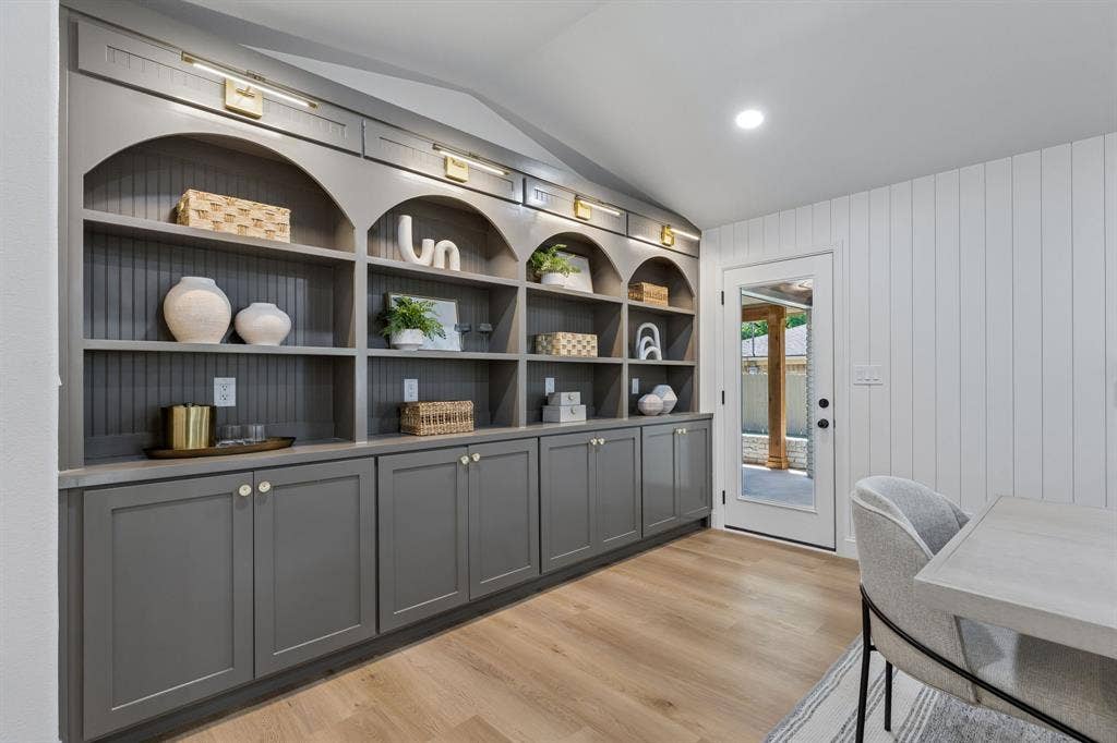 12124 Dixfield Drive, Dallas, TX 75218 Vaulted ceiling room featuring extensive built-in shelving with arched details, lower cabinetry with brass hardware, wood-finish flooring, and a glass-paneled door