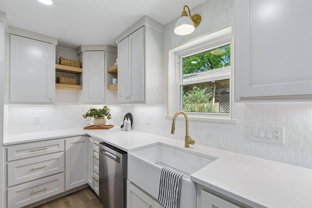 12124 Dixfield Drive, Dallas, TX 75218 White cabinetry with gold-tone hardware, white countertops, built-in stainless steel dishwasher, and a white farmhouse sink with a gold-tone faucet