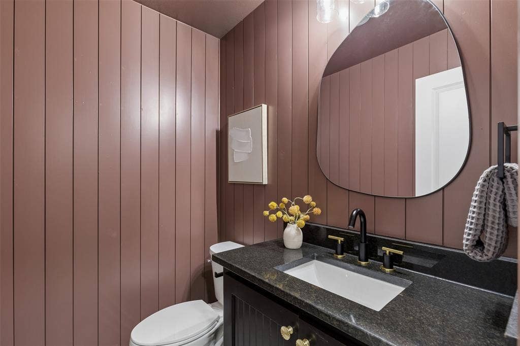 12124 Dixfield Drive, Dallas, TX 75218 Powder room featuring vertical shiplap paneling in warm mauve tones, a curved organic mirror with integrated lighting, dark stone countertop with undermount sink basin, matte black faucet hardware, white porcelain toilet, and recessed wall-mounted medicine cabinet