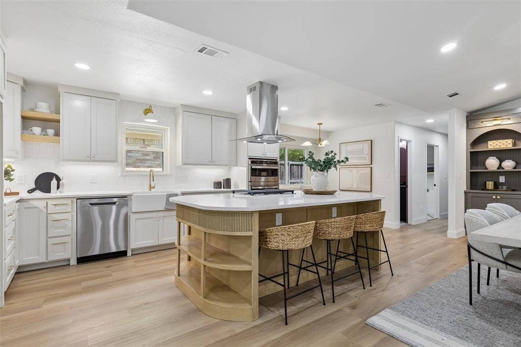 12124 Dixfield Drive, Dallas, TX 75218 Gourmet kitchen with wood-finish flooring, white cabinetry, a large kitchen island with built-in shelving, stainless steel appliances, and recessed lighting
