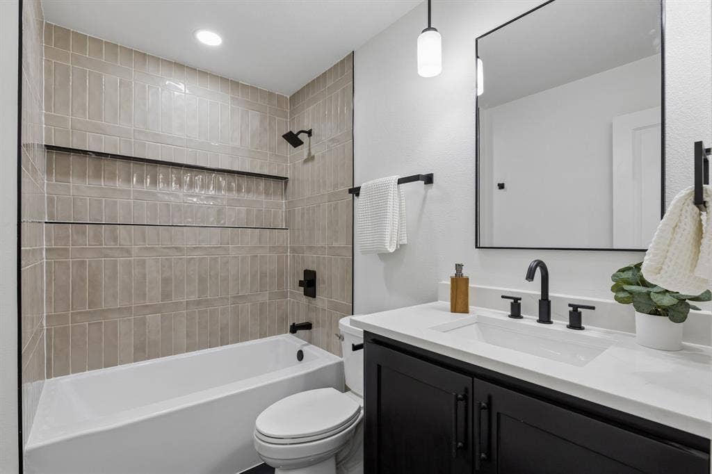 12124 Dixfield Drive, Dallas, TX 75218 Bathtub and shower combination with vertical tile surround and built-in shelving, black vanity with an integrated sink and white countertop, black framed mirror, and matte black fixtures