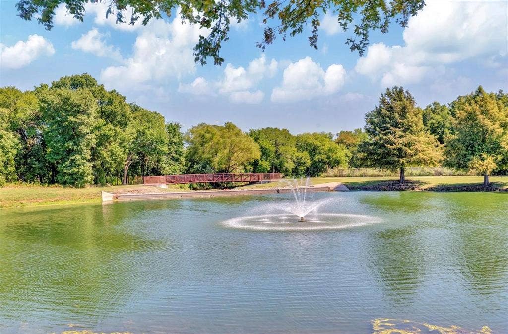 1601 Long Prairie Court, Allen, TX 75002 Pond with a central fountain, surrounded by mature trees, a grassy bank, and a pedestrian bridge