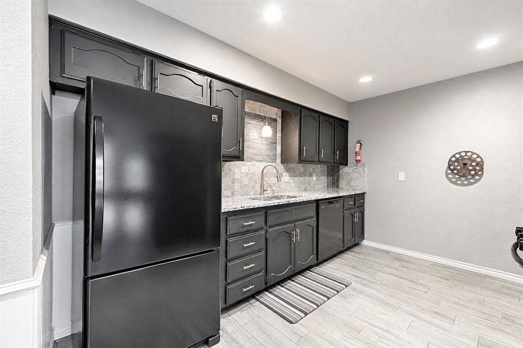 1223 Sequoia Drive, DeSoto, TX 75115 Black cabinetry with decorative molding, light-toned tile backsplash, granite-style countertops, integrated stainless-steel sink, and wood-finish flooring