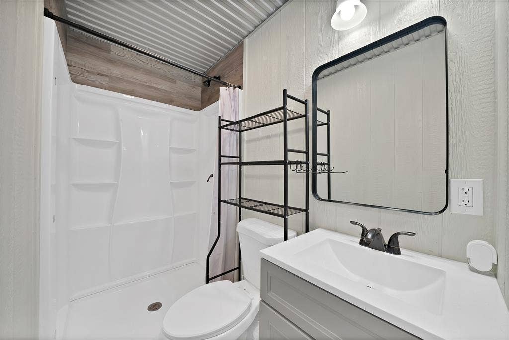 Walk-in shower with integrated shelving, gray vanity with white countertop and integrated sink, black-framed mirror, and wood-finish wall accent