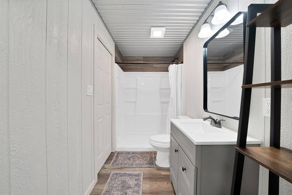 White paneled walls, corrugated metal ceiling, wood-finish flooring, full bath with shower, and a vanity with an integrated sink
