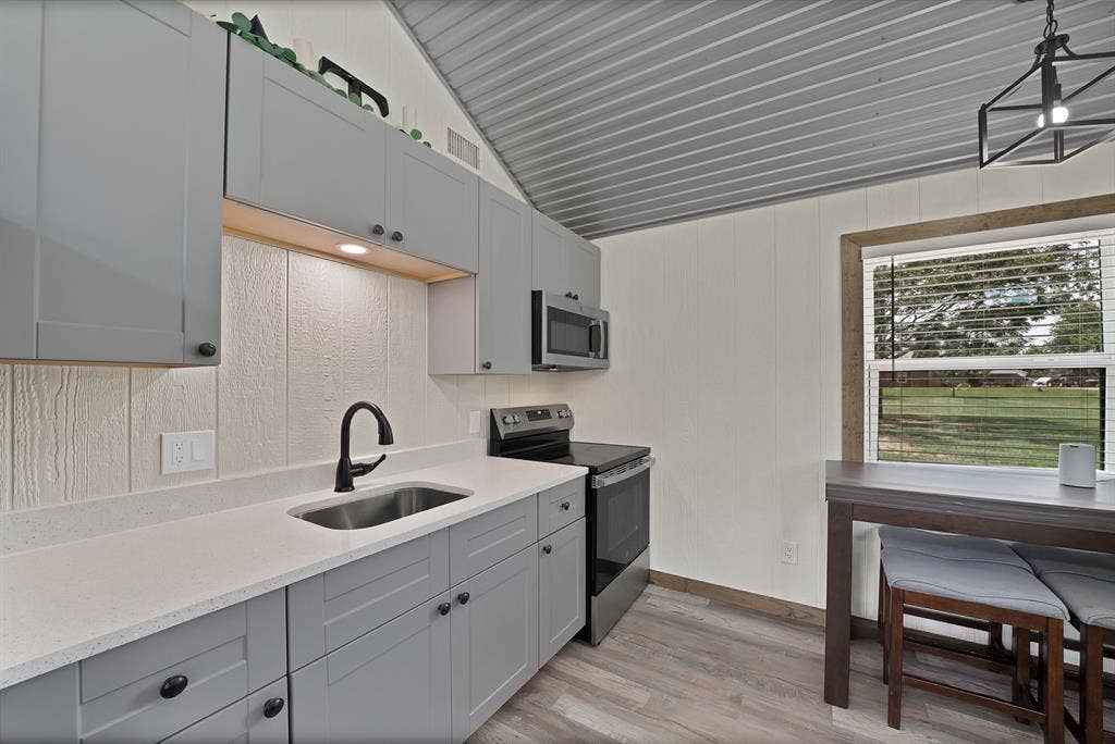 Light gray cabinetry with matte black hardware, under-cabinet lighting, stainless steel sink with a matte black faucet, and wood-finish flooring