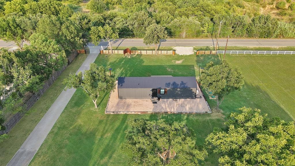 Expansive equestrian property featuring a modern stable structure with dark roof and concrete foundation, manicured pasture grounds, white rail fencing, mature tree-lined perimeter, and a paved driveway providing direct access to the facilities