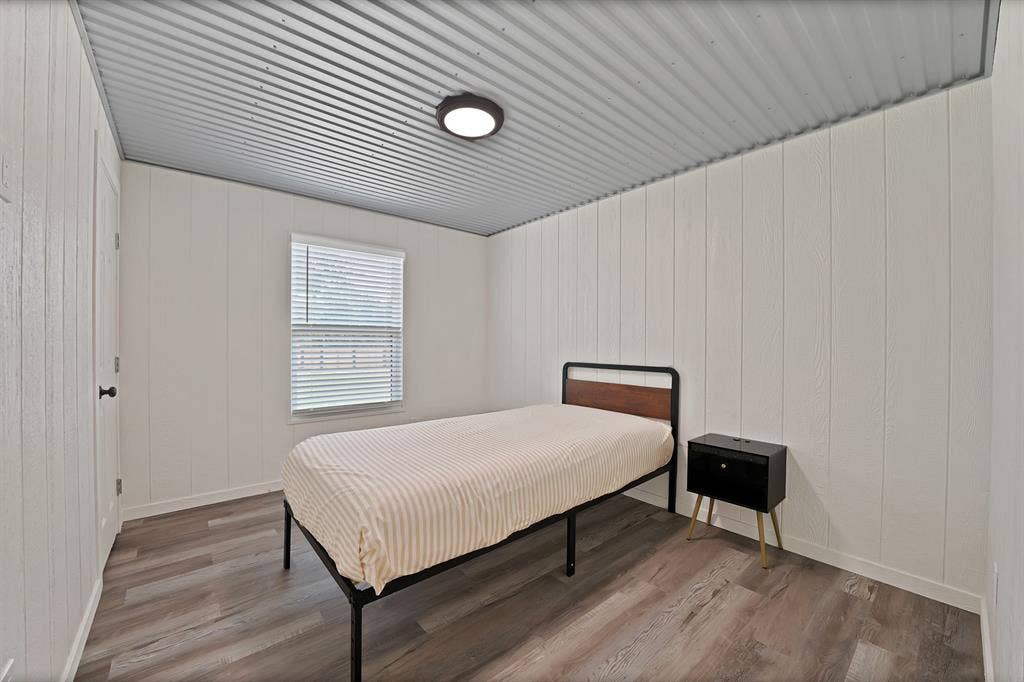 Corrugated metal ceiling, wood-finish flooring, white vertical plank walls, and a window with blinds