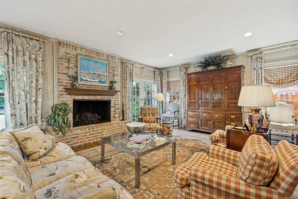 3418 Dartmouth Avenue, Highland Park, TX 75205 Elegant living room showcasing a brick fireplace with decorative mantel, hardwood flooring, layered drapery with coordinating patterns, recessed ceiling lighting, and substantial wood cabinetry