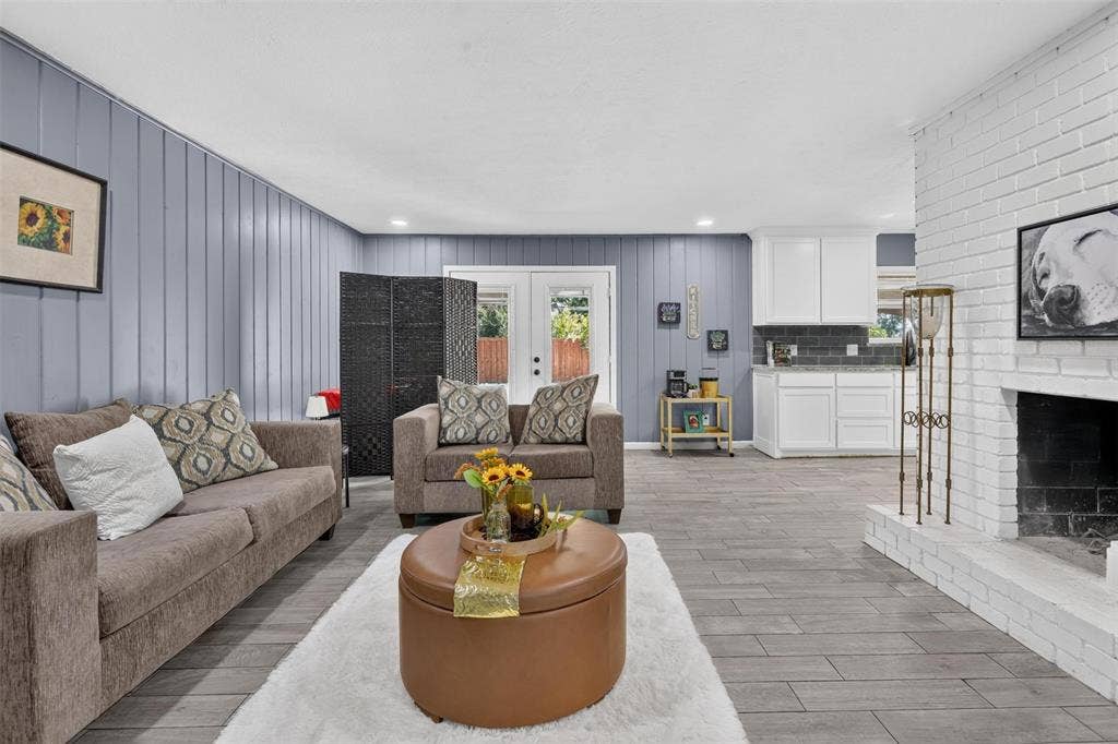 8117 Rush Street, Benbrook, TX 76116 Open-concept living space featuring gray wood-finish flooring throughout, neutral upholstered seating, a round cognac-leather ottoman, and a fireplace with white shiplap surround