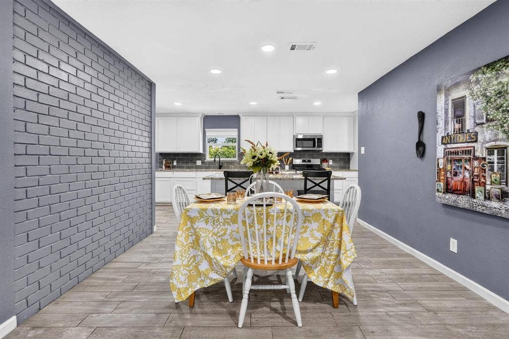 8117 Rush Street, Benbrook, TX 76116 Open-concept dining and kitchen area featuring recessed ceiling lighting, gray painted accent walls, white cabinetry with stainless steel appliances, wood-finish flooring, and a brick accent wall with modern geometric detailing