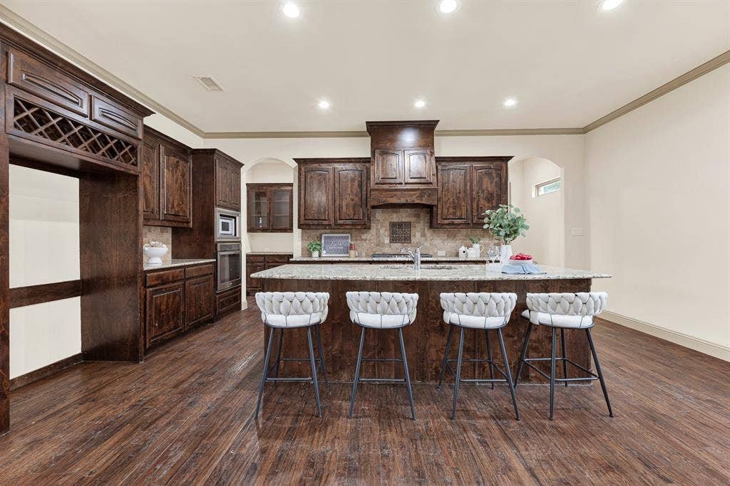 7013 BROOK FOREST Circle, Plano, TX 75024 A striking carved wood range hood and rich espresso cabinetry anchor the back wall under crisp ceiling downlights. Imagine rolling out homemade pizza dough across the expansive granite island while family gathers around to help.