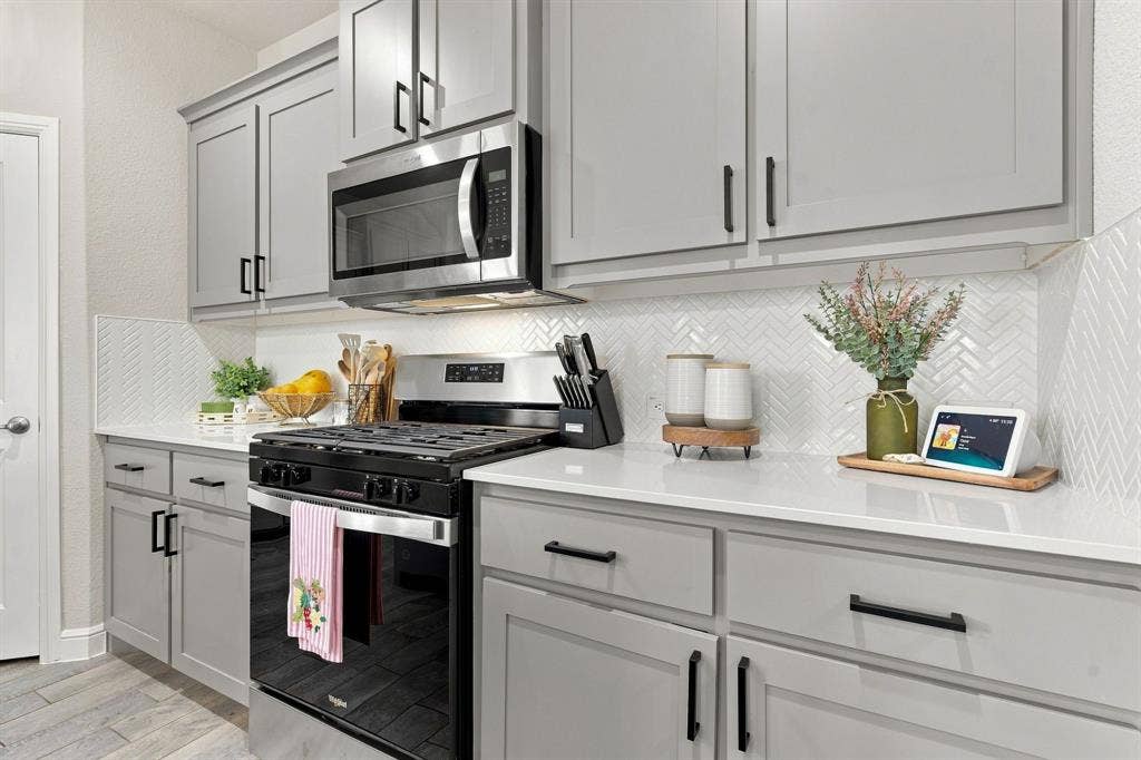 Gray shaker-style cabinetry with black hardware, white countertops, herringbone tile backsplash, stainless steel microwave, and a black gas range