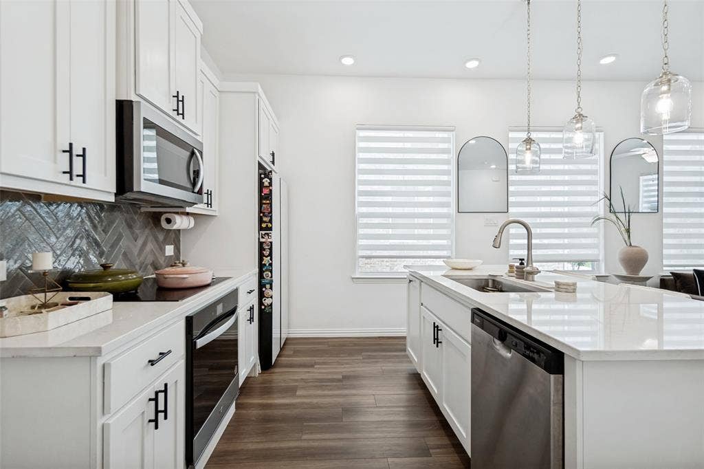 7536 Laburnam Road, Dallas, TX 75228 White cabinetry with black hardware, wood-finish flooring, stainless steel appliances, herringbone tile backsplash, and a kitchen island with an integrated sink