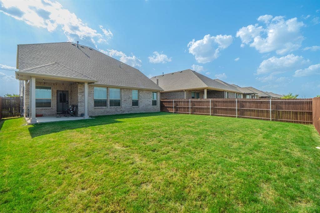 Expansive lawn area with a wood privacy fence, brick exterior, covered patio, and multiple windows