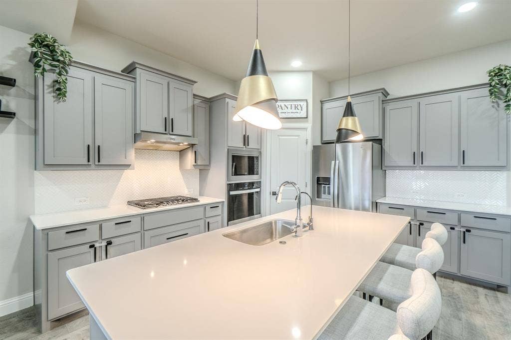 Spacious kitchen featuring an island with an undermount sink, light gray cabinetry, stainless steel appliances, a gas cooktop, and herringbone tile backsplash