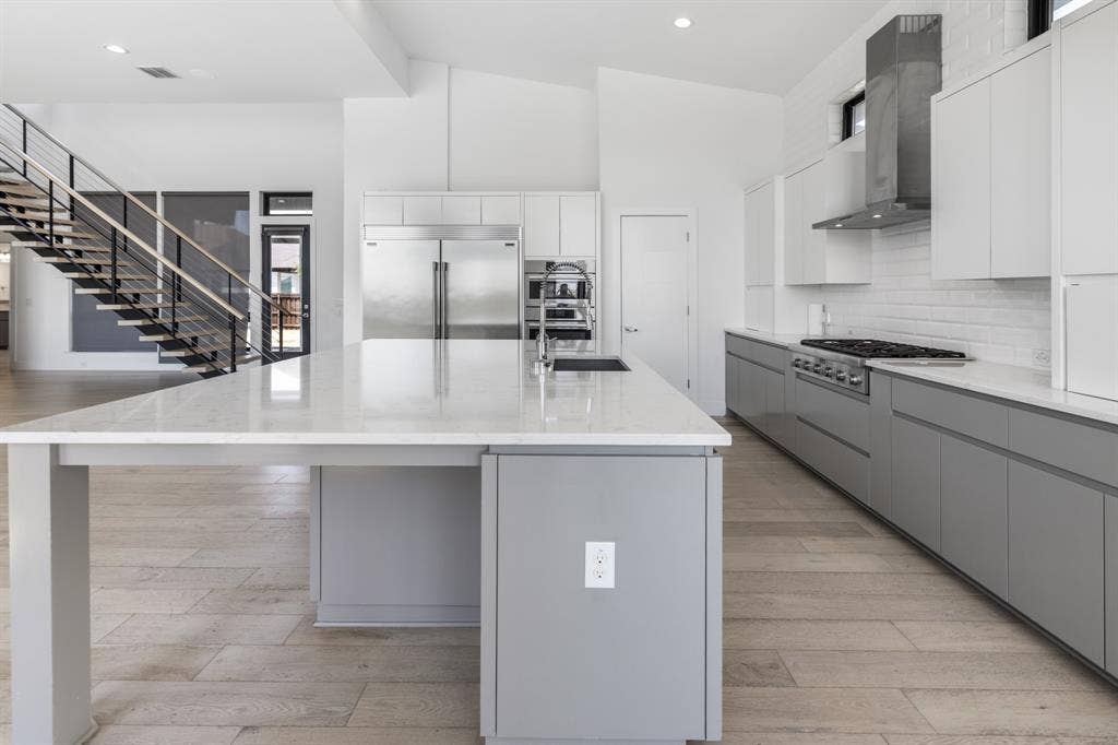 1321 Black Walnut Lane, Arlington, TX 76005 Modern kitchen featuring a large center island with waterfall countertop edge, stainless steel appliances including a built-in refrigerator and range hood, white cabinetry with integrated hardware, quartz countertops, and wood-finish flooring throughout
