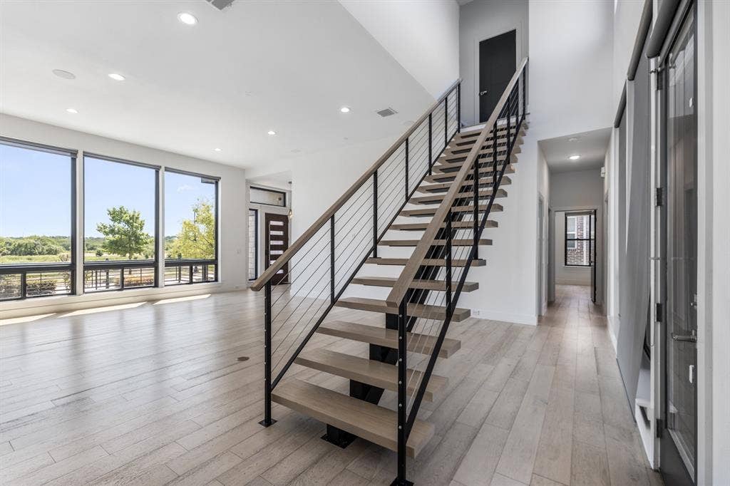 1321 Black Walnut Lane, Arlington, TX 76005 Modern open-concept space featuring a floating staircase with black metal railings and wood-finish treads, light wood-finish flooring throughout, floor-to-ceiling windows with expansive natural light, and recessed ceiling lighting