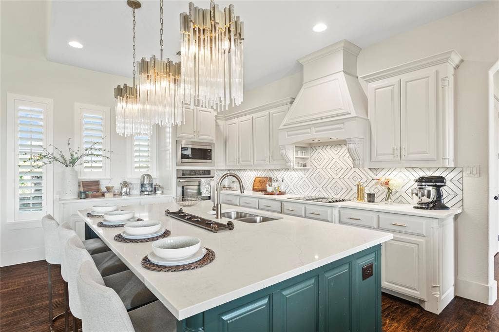 623 Calm Water Cove, Princeton, TX 75407 Kitchen with a teal island, white perimeter cabinetry, a white range hood, a patterned tile backsplash, and three tiered glass and brass chandeliers