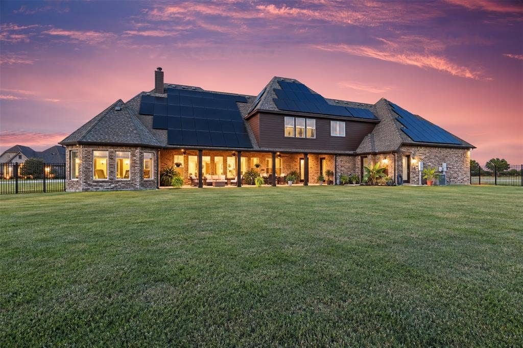 623 Calm Water Cove, Princeton, TX 75407 Expansive rear facade featuring brick and siding, multiple solar panel arrays, a covered patio, and a large grassy yard enclosed by black fencing