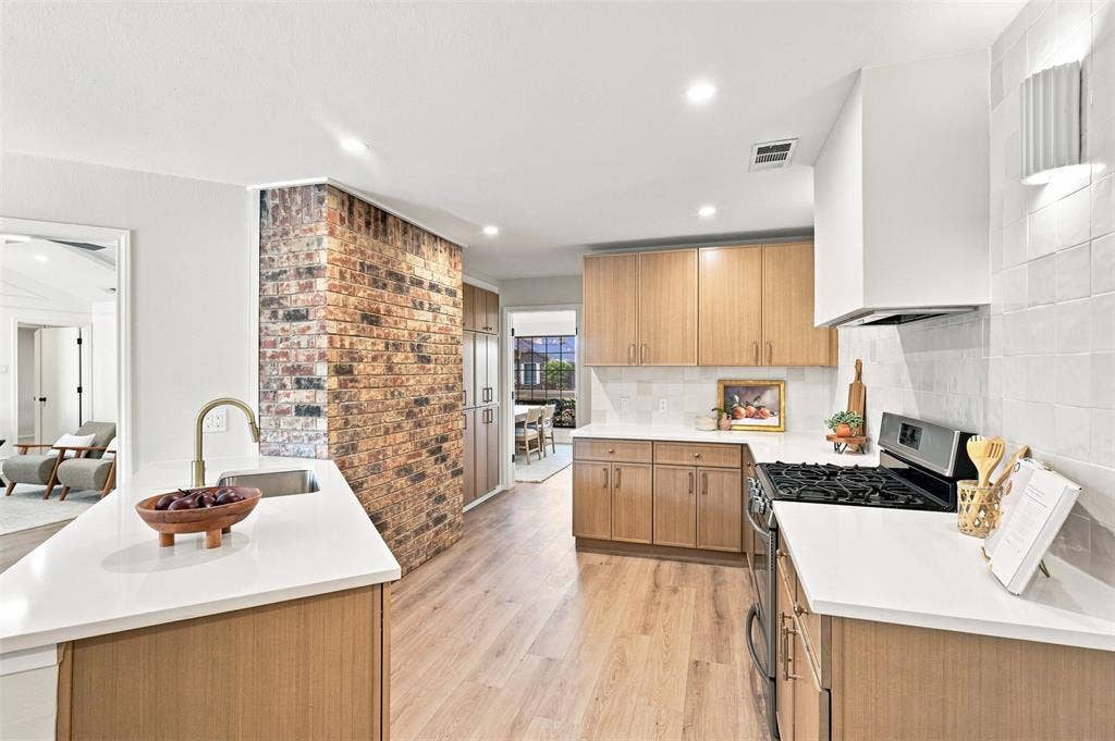 2411 Aspen Street, Richardson, TX 75082 Modern kitchen featuring dual islands with wood-finish cabinetry and white quartz countertops, stainless steel gas cooktop, custom hood ventilation, exposed brick accent wall, and recessed ceiling lighting throughout the open layout with hardwood flooring