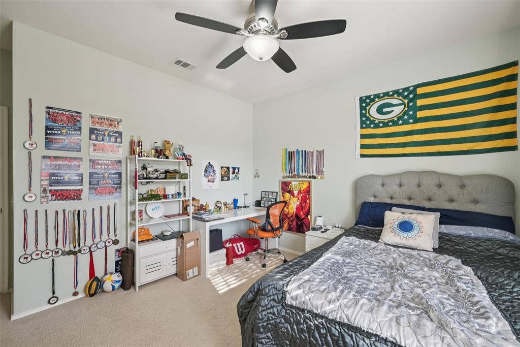 Light-colored walls, ceiling fan with integrated lighting, built-in shelving, desk with ample workspace, and soft carpet flooring