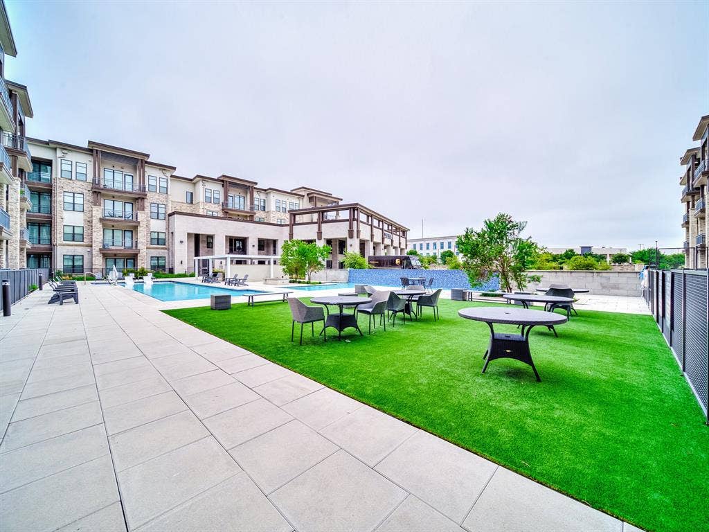 2700 Old Denton Road, #2212, Carrollton, TX 75007 Expansive outdoor space featuring a rectangular swimming pool, paver stone hardscaping, artificial turf lawn, and a multi-story building exterior with stone and siding accents