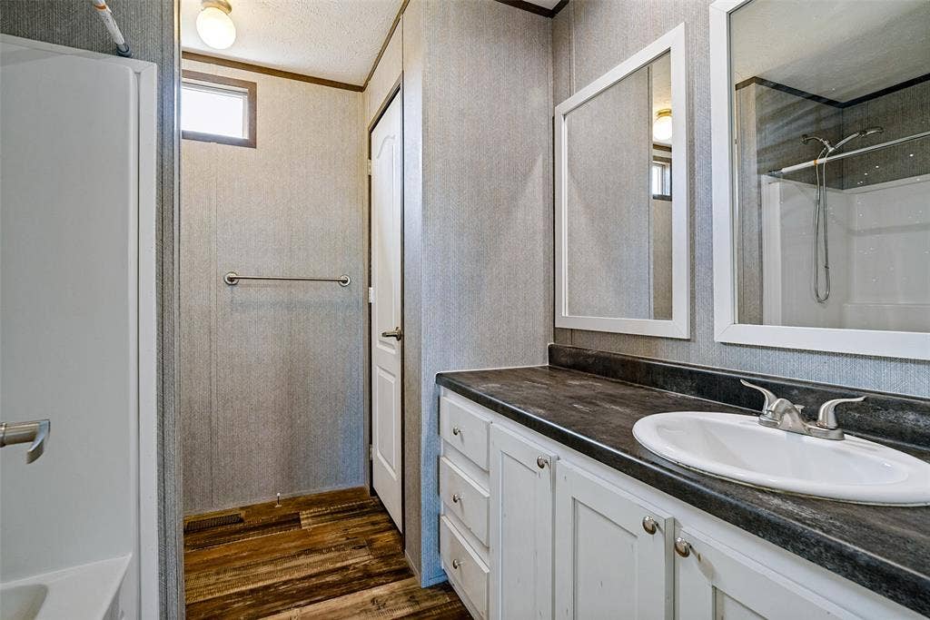 4415 Woodland Drive, Granbury, TX 76048 Wood-finish flooring, white vanity with cabinet storage, countertop with integrated sink, framed mirrors, and a built-in shower