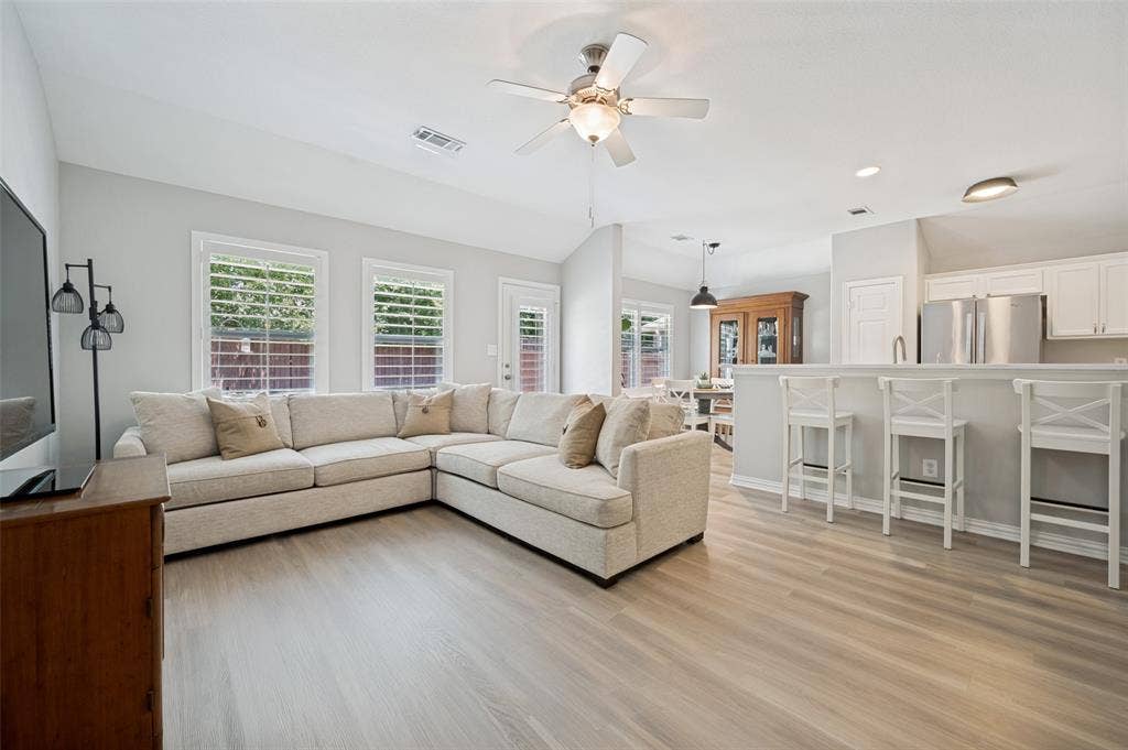 3707 Redwood Circle, Melissa, TX 75454 Open-concept living and kitchen area featuring a cream upholstered sectional sofa, wood-finish flooring throughout, stainless steel appliances, white cabinetry, and abundant natural light from multiple windows with plantation shutters