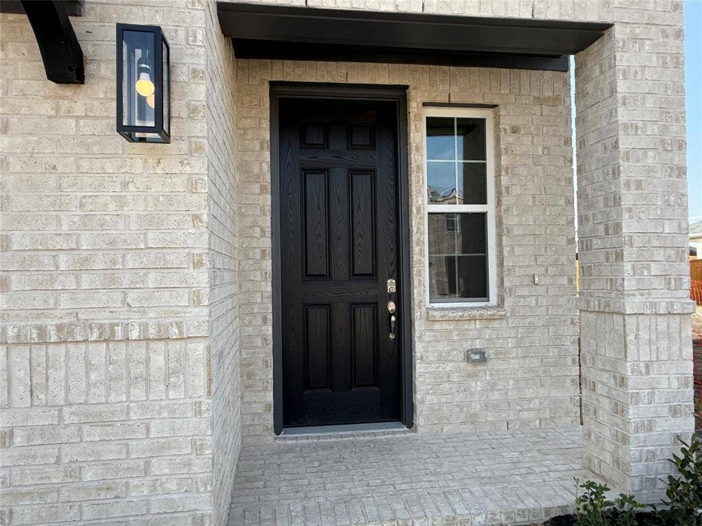 Light-toned brick exterior with a dark-paneled entry door, a single-pane window with grid inserts, an exterior wall lantern, and a covered porch
