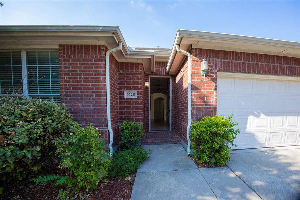 5728 Calloway Drive, McKinney, TX 75070 Brick exterior with an attached garage, featuring a concrete walkway, mature landscaping, and an exterior light fixture