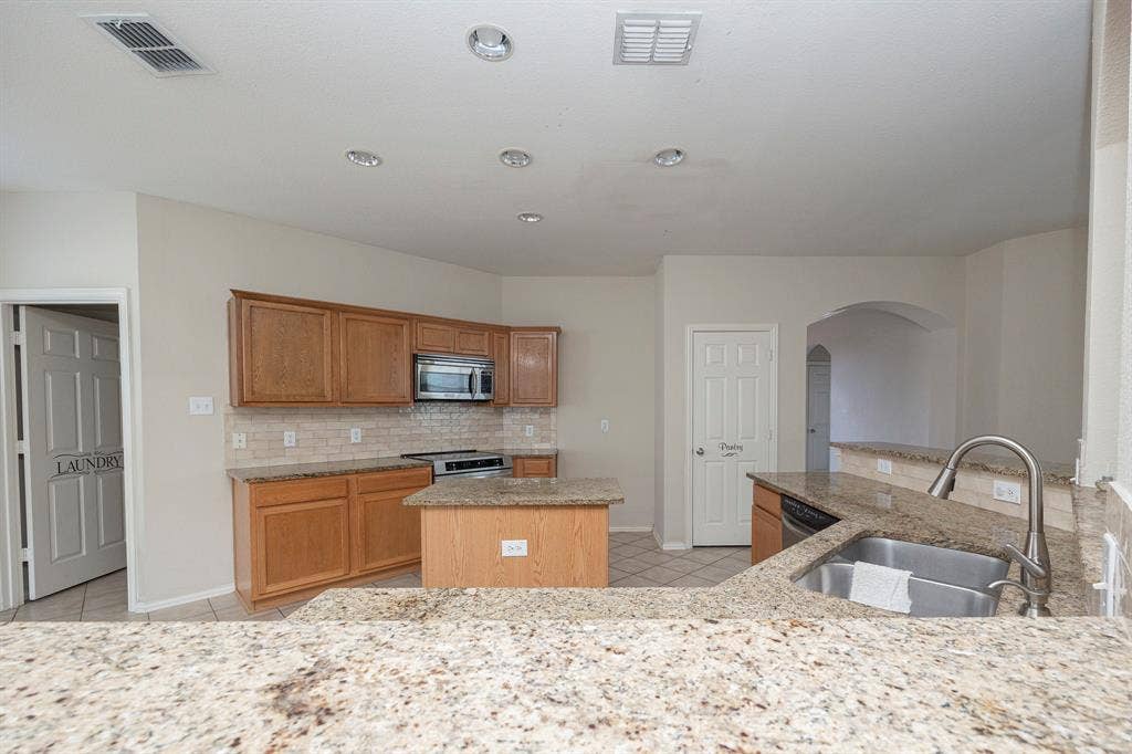 5728 Calloway Drive, McKinney, TX 75070 Spacious kitchen featuring warm wood cabinetry, stainless steel appliances including a built-in microwave, a large center island with seating, speckled stone countertops, and a tile backsplash