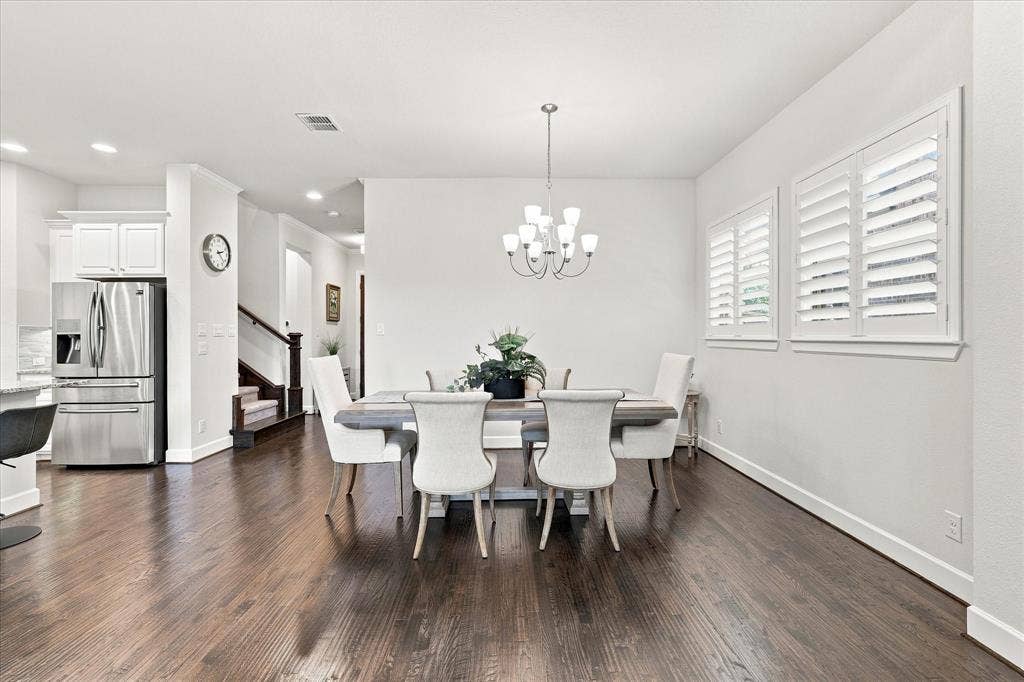 Elegant dining space featuring a wood-finish plank floor, a round pedestal table with upholstered chairs, a multi-arm chandelier, and plantation shutters flanking the windows