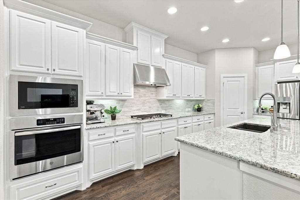 White shaker cabinetry with crown molding, stainless steel built-in appliances, light-toned stone countertops, a tile backsplash, and wood-finish flooring