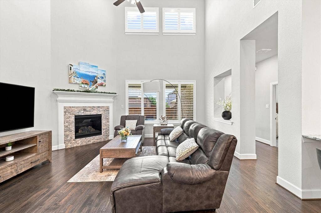 Open-concept living area featuring soaring ceilings with ceiling fan, hardwood flooring throughout, and a brick fireplace with surround
