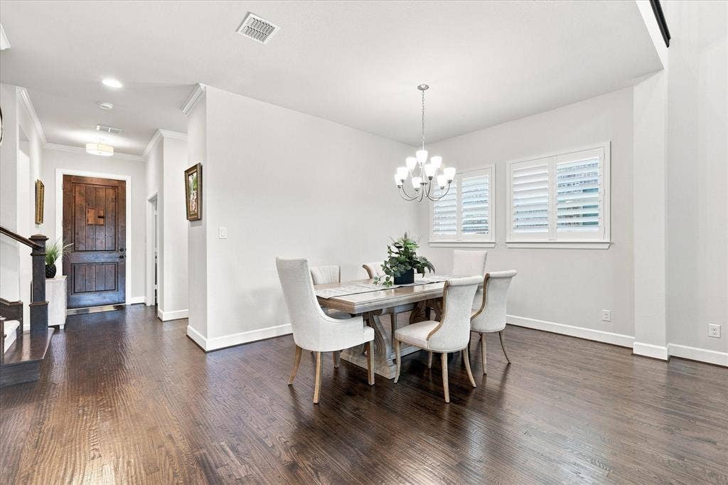 Elegant dining room showcasing rich wood-finish flooring, a chandelier pendant fixture, and abundant natural light from multiple shuttered windows