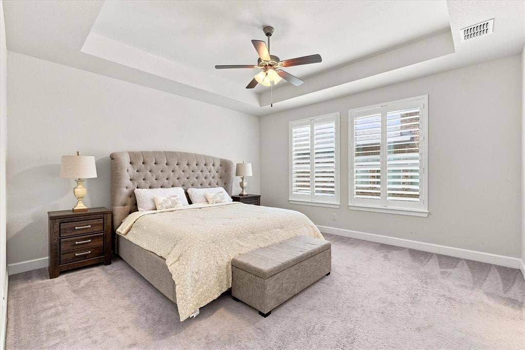 Tray ceiling with a ceiling fan, two windows with plantation shutters, and light-toned carpet flooring