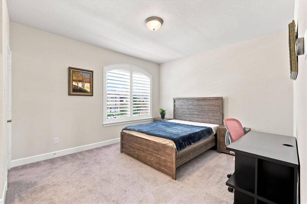 Bright secondary bedroom featuring neutral carpeting, wood-finish bed frame with upholstered headboard, plantation shutters with light-filtering louvers, recessed ceiling lighting, and dedicated workspace with black desk and chair