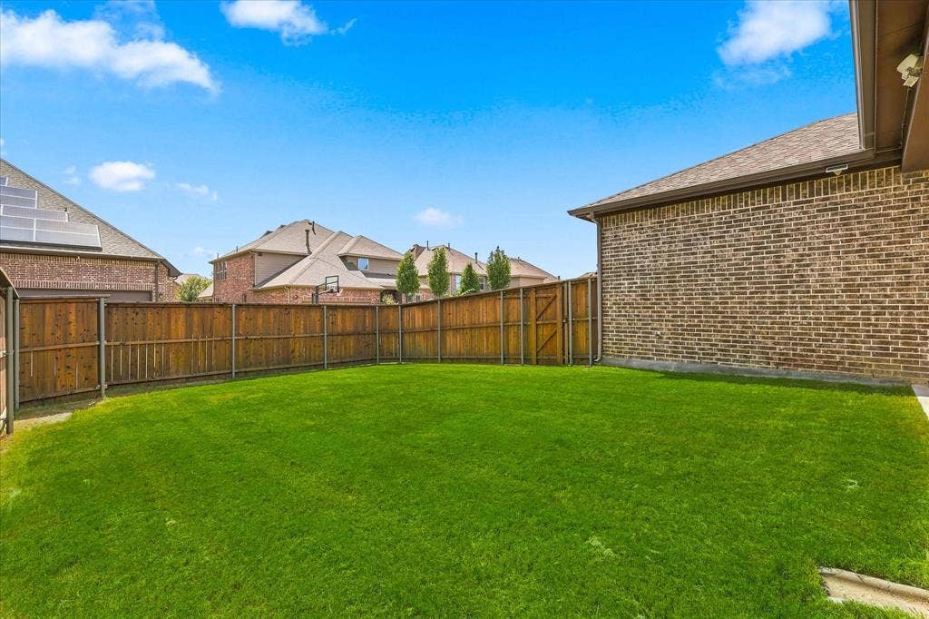 Expansive backyard featuring a pristine manicured lawn bordered by wood-finish fencing, with mature landscaping and brick-constructed homes visible beyond the perimeter